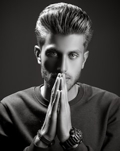 A grayscale portrait of a man with styled hair, looking intently at the camera with his hands placed together in front of his face. He wears a textured sweater and several bracelets on one wrist. The lighting is dramatic, creating strong shadows and highlights to emphasize his facial features.