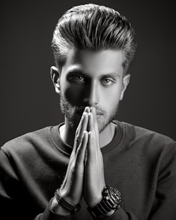 A grayscale portrait of a man with styled hair, looking intently at the camera with his hands placed together in front of his face. He wears a textured sweater and several bracelets on one wrist. The lighting is dramatic, creating strong shadows and highlights to emphasize his facial features.