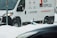 A white delivery van with 'ALPI EXPRESS' branding is parked on a snowy street. Snow is falling, creating a wintry scene, and a partially buried scooter can be seen in the snow near the van.