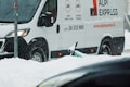 A white delivery van with 'ALPI EXPRESS' branding is parked on a snowy street. Snow is falling, creating a wintry scene, and a partially buried scooter can be seen in the snow near the van.