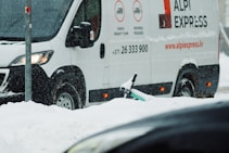 A white delivery van with 'ALPI EXPRESS' branding is parked on a snowy street. Snow is falling, creating a wintry scene, and a partially buried scooter can be seen in the snow near the van.
