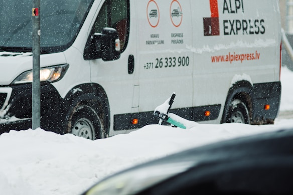 A white delivery van with 'ALPI EXPRESS' branding is parked on a snowy street. Snow is falling, creating a wintry scene, and a partially buried scooter can be seen in the snow near the van.