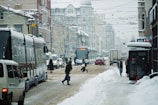 A snowy urban street with a tram and several people walking across the slushy road. The street is lined with parked vehicles and is bustling with traffic. Snow covers the street and buildings, and overhead tram cables are visible.