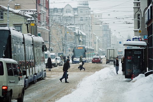 A snowy urban street with a tram and several people walking across the slushy road. The street is lined with parked vehicles and is bustling with traffic. Snow covers the street and buildings, and overhead tram cables are visible.