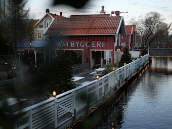 A rustic red building with the word 'B&Aring;TBYGGERI' written on it is situated by the water, surrounded by Christmas trees and illuminated with small lights. The scene captures a charming, wintry village ambiance with a wooden walkway lined by a white fence, leading up to the structure. The area is tranquil with bare trees in the background and other similar styled buildings nearby.