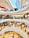 A modern, multi-level shopping mall with numerous stores and people walking around. The architecture features large, curved balconies and a high ceiling with skylights. Shoppers are visible on each floor, browsing through various shops and interacting with their surroundings.