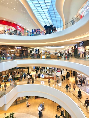 A modern, multi-level shopping mall with numerous stores and people walking around. The architecture features large, curved balconies and a high ceiling with skylights. Shoppers are visible on each floor, browsing through various shops and interacting with their surroundings.