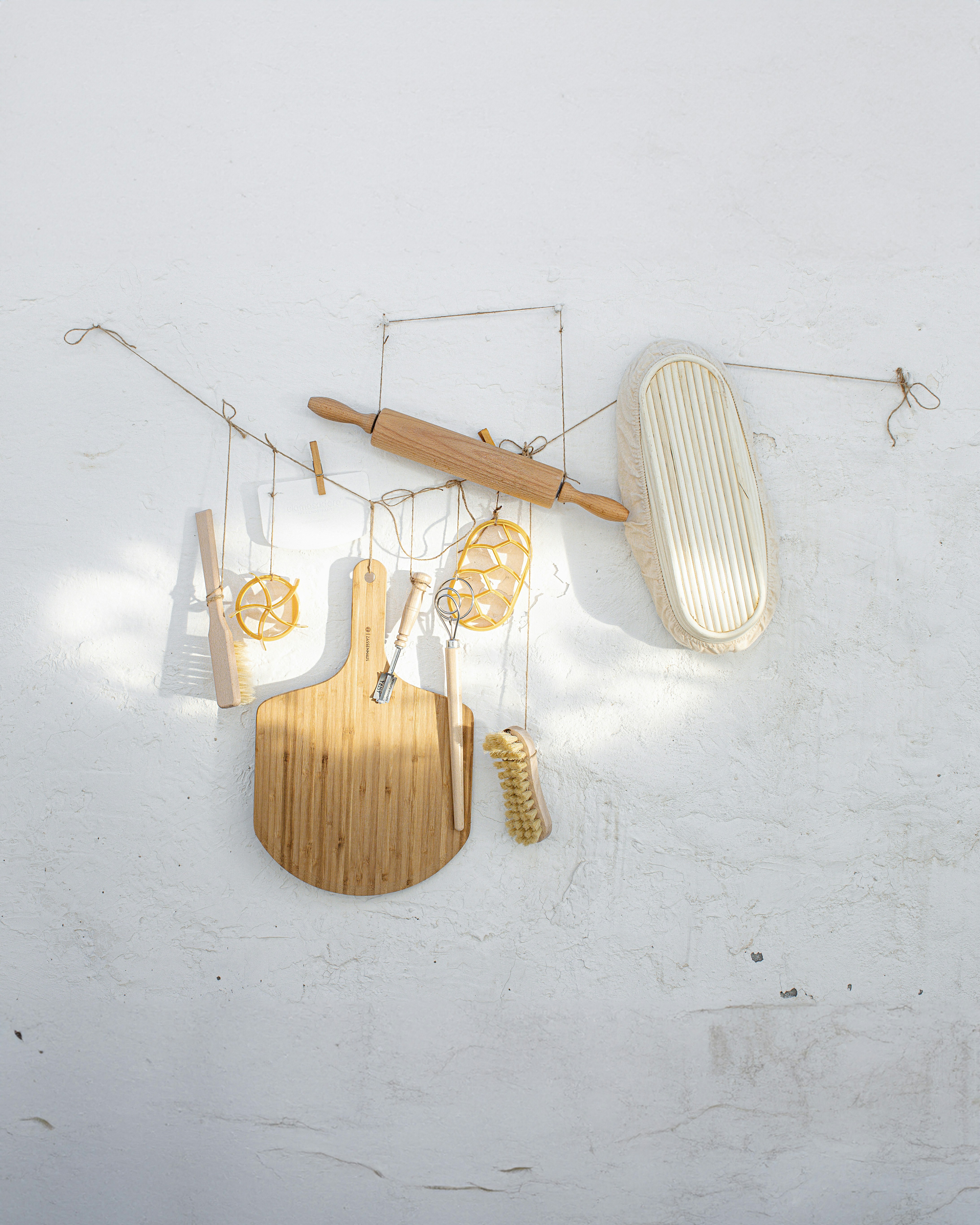 A variety of kitchen utensils are artistically arranged against a textured white wall. The collection includes a wooden rolling pin, a cutting board, a whisk, a brush, and a few other tools, all suspended with delicate strings.