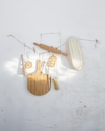 A variety of kitchen utensils are artistically arranged against a textured white wall. The collection includes a wooden rolling pin, a cutting board, a whisk, a brush, and a few other tools, all suspended with delicate strings.