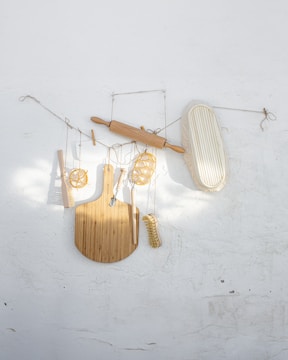 A variety of kitchen utensils are artistically arranged against a textured white wall. The collection includes a wooden rolling pin, a cutting board, a whisk, a brush, and a few other tools, all suspended with delicate strings.