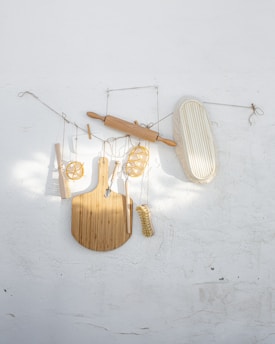 A variety of kitchen utensils are artistically arranged against a textured white wall. The collection includes a wooden rolling pin, a cutting board, a whisk, a brush, and a few other tools, all suspended with delicate strings.