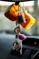 A pair of fuzzy yellow dice and a small jar hang from a car rearview mirror, accompanied by a set of keys and various keychains including a plush keychain. The objects are hanging against a blurred interior car background.