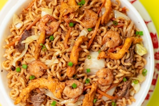 A close-up view of a bowl filled with stir-fried noodles, featuring shrimp, sliced vegetables, and chunks of meat, garnished with chopped green onions. The noodles are intertwined, giving a textured look, while the vegetables add color and variety to the dish.