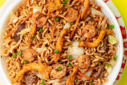 A close-up view of a bowl filled with stir-fried noodles, featuring shrimp, sliced vegetables, and chunks of meat, garnished with chopped green onions. The noodles are intertwined, giving a textured look, while the vegetables add color and variety to the dish.