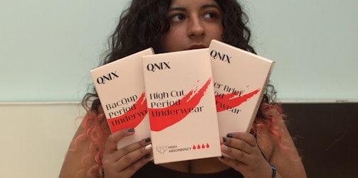 A person with long dark curly hair is holding three boxes labeled QNIX. The packages are of menstrual underwear with labels such as 'Bacoup Period Underwear' and 'High Cut Period Underwear'. The background is plain, and the person has their gaze directed away from the camera.