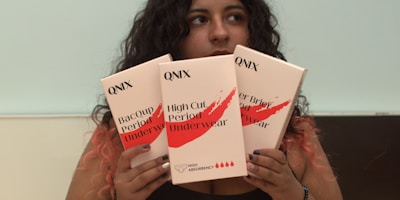 A person with long dark curly hair is holding three boxes labeled QNIX. The packages are of menstrual underwear with labels such as 'Bacoup Period Underwear' and 'High Cut Period Underwear'. The background is plain, and the person has their gaze directed away from the camera.