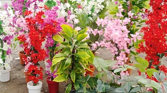 A collection of vibrant, artificial flower arrangements in pots, featuring vivid red, pink, purple, and white hues. The flowers are mixed with green leaves and resemble a garden display on a dirt ground.