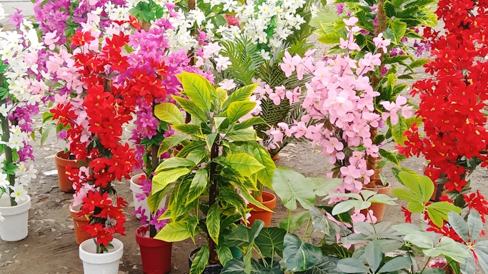 A collection of vibrant, artificial flower arrangements in pots, featuring vivid red, pink, purple, and white hues. The flowers are mixed with green leaves and resemble a garden display on a dirt ground.