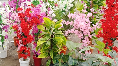 A collection of vibrant, artificial flower arrangements in pots, featuring vivid red, pink, purple, and white hues. The flowers are mixed with green leaves and resemble a garden display on a dirt ground.