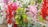 A collection of vibrant, artificial flower arrangements in pots, featuring vivid red, pink, purple, and white hues. The flowers are mixed with green leaves and resemble a garden display on a dirt ground.