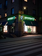 A street scene at dusk featuring a pharmacy located at the corner of a building. The pharmacy has a bright neon sign with the word 'APOTEK' and heart symbols, glowing in green. The front windows display illuminated shelves with products. A pedestrian crossing is visible in the foreground under soft street lighting.