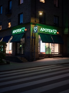 A street scene at dusk featuring a pharmacy located at the corner of a building. The pharmacy has a bright neon sign with the word 'APOTEK' and heart symbols, glowing in green. The front windows display illuminated shelves with products. A pedestrian crossing is visible in the foreground under soft street lighting.