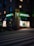 A street scene at dusk featuring a pharmacy located at the corner of a building. The pharmacy has a bright neon sign with the word 'APOTEK' and heart symbols, glowing in green. The front windows display illuminated shelves with products. A pedestrian crossing is visible in the foreground under soft street lighting.