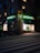 A street scene at dusk featuring a pharmacy located at the corner of a building. The pharmacy has a bright neon sign with the word 'APOTEK' and heart symbols, glowing in green. The front windows display illuminated shelves with products. A pedestrian crossing is visible in the foreground under soft street lighting.