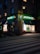 A street scene at dusk featuring a pharmacy located at the corner of a building. The pharmacy has a bright neon sign with the word 'APOTEK' and heart symbols, glowing in green. The front windows display illuminated shelves with products. A pedestrian crossing is visible in the foreground under soft street lighting.