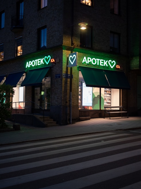 A street scene at dusk featuring a pharmacy located at the corner of a building. The pharmacy has a bright neon sign with the word 'APOTEK' and heart symbols, glowing in green. The front windows display illuminated shelves with products. A pedestrian crossing is visible in the foreground under soft street lighting.