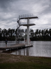 A tall diving platform stands beside a body of water. The platform has three levels with a spiral staircase on one side. There are three people near the platform, two standing on the dock and one walking away. The scene is set under a cloudy sky with a row of trees visible in the background.