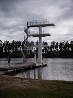 A tall diving platform stands beside a body of water. The platform has three levels with a spiral staircase on one side. There are three people near the platform, two standing on the dock and one walking away. The scene is set under a cloudy sky with a row of trees visible in the background.