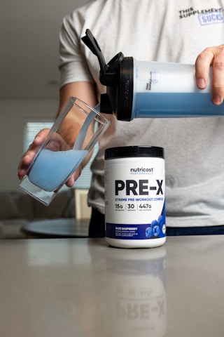 A person pours blue liquid from a shaker bottle into a glass. Next to the glass is a container labeled 'PRE-X' which appears to be a pre-workout supplement. The scene suggests a focus on fitness or nutrition.