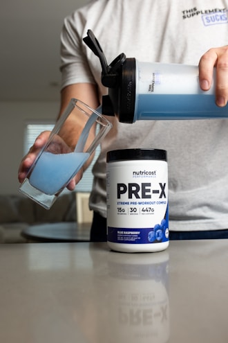 A person pours blue liquid from a shaker bottle into a glass. Next to the glass is a container labeled 'PRE-X' which appears to be a pre-workout supplement. The scene suggests a focus on fitness or nutrition.