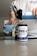 A person pours blue liquid from a shaker bottle into a glass. Next to the glass is a container labeled 'PRE-X' which appears to be a pre-workout supplement. The scene suggests a focus on fitness or nutrition.