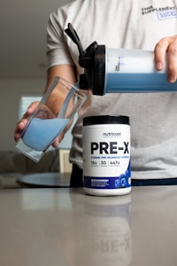 A person pours blue liquid from a shaker bottle into a glass. Next to the glass is a container labeled 'PRE-X' which appears to be a pre-workout supplement. The scene suggests a focus on fitness or nutrition.