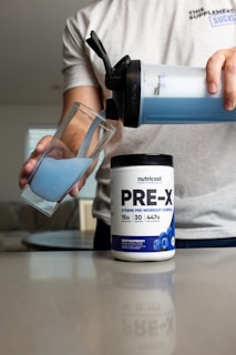 A person pours blue liquid from a shaker bottle into a glass. Next to the glass is a container labeled 'PRE-X' which appears to be a pre-workout supplement. The scene suggests a focus on fitness or nutrition.