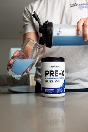 A person pours blue liquid from a shaker bottle into a glass. Next to the glass is a container labeled 'PRE-X' which appears to be a pre-workout supplement. The scene suggests a focus on fitness or nutrition.