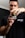 A person wearing a black t-shirt is holding a container labeled 'Psychotic' by Insane Labz. The focus is on the container, with the person's face slightly blurred in the background. The t-shirt has a logo that includes the text 'This Supplement Sucks'. The setting appears to be indoors with neutral-colored walls.