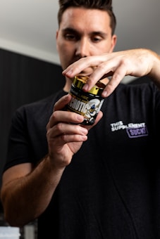 A person wearing a black t-shirt is holding a container labeled 'Psychotic' by Insane Labz. The focus is on the container, with the person's face slightly blurred in the background. The t-shirt has a logo that includes the text 'This Supplement Sucks'. The setting appears to be indoors with neutral-colored walls.