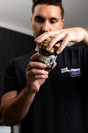 A person wearing a black t-shirt is holding a container labeled 'Psychotic' by Insane Labz. The focus is on the container, with the person's face slightly blurred in the background. The t-shirt has a logo that includes the text 'This Supplement Sucks'. The setting appears to be indoors with neutral-colored walls.