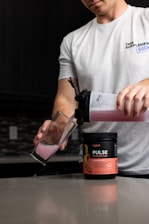A person is pouring a pink liquid from a shaker bottle into a glass. The shaker bottle is positioned over a container labeled 'Pulse Pre-Workout Drink' with a strawberry kiwi flavor. The person is wearing a white t-shirt and is in a kitchen setting with a dark backsplash and countertop.