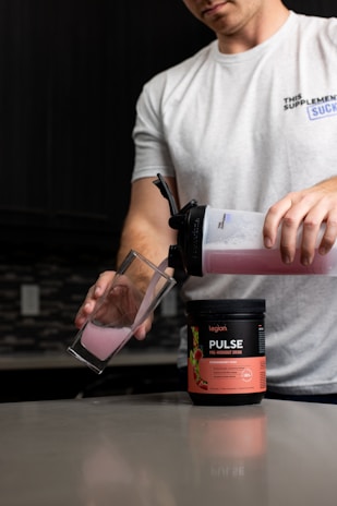 A person is pouring a pink liquid from a shaker bottle into a glass. The shaker bottle is positioned over a container labeled 'Pulse Pre-Workout Drink' with a strawberry kiwi flavor. The person is wearing a white t-shirt and is in a kitchen setting with a dark backsplash and countertop.