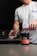 A person is pouring a pink liquid from a shaker bottle into a glass. The shaker bottle is positioned over a container labeled 'Pulse Pre-Workout Drink' with a strawberry kiwi flavor. The person is wearing a white t-shirt and is in a kitchen setting with a dark backsplash and countertop.