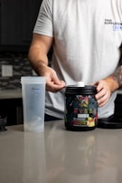 A person with a tattooed arm is wearing a white t-shirt and is preparing a workout supplement. The person holds a scoop over an open container of pre-workout powder labeled 'RYSE Godzilla Strawberry Kiwi'. Beside the container is a clear plastic shaker bottle on a kitchen counter.