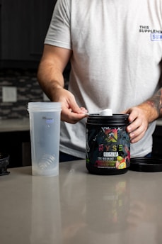 A person with a tattooed arm is wearing a white t-shirt and is preparing a workout supplement. The person holds a scoop over an open container of pre-workout powder labeled 'RYSE Godzilla Strawberry Kiwi'. Beside the container is a clear plastic shaker bottle on a kitchen counter.