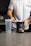 A person with a tattooed arm is wearing a white t-shirt and is preparing a workout supplement. The person holds a scoop over an open container of pre-workout powder labeled 'RYSE Godzilla Strawberry Kiwi'. Beside the container is a clear plastic shaker bottle on a kitchen counter.