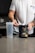 A person with a tattooed arm is wearing a white t-shirt and is preparing a workout supplement. The person holds a scoop over an open container of pre-workout powder labeled 'RYSE Godzilla Strawberry Kiwi'. Beside the container is a clear plastic shaker bottle on a kitchen counter.