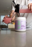 A person is pouring a pink-colored drink from a shaker bottle into a glass. A container labeled 'Thavage Pre-Workout' is placed on a countertop in front of the person. The surroundings appear to be an indoor setting with soft lighting and blurred background elements.