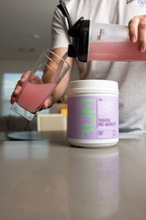 A person is pouring a pink-colored drink from a shaker bottle into a glass. A container labeled 'Thavage Pre-Workout' is placed on a countertop in front of the person. The surroundings appear to be an indoor setting with soft lighting and blurred background elements.